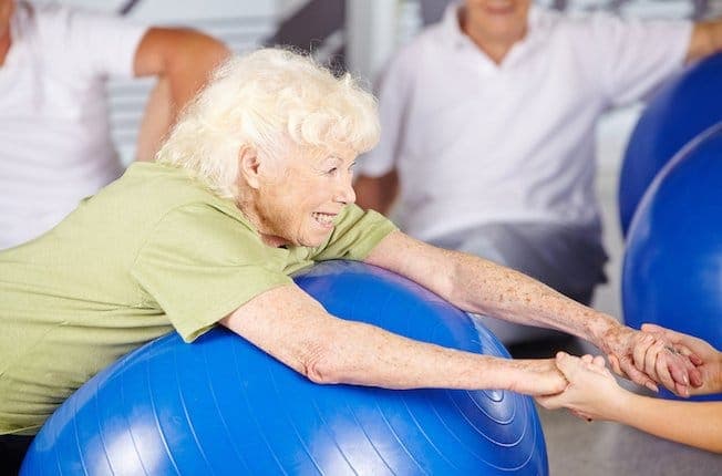 Therapeutin hält die Hände einer lächelnden älteren Patientin, die sich beim Gleichgewichtstraining auf einen Gymnastikball stützt