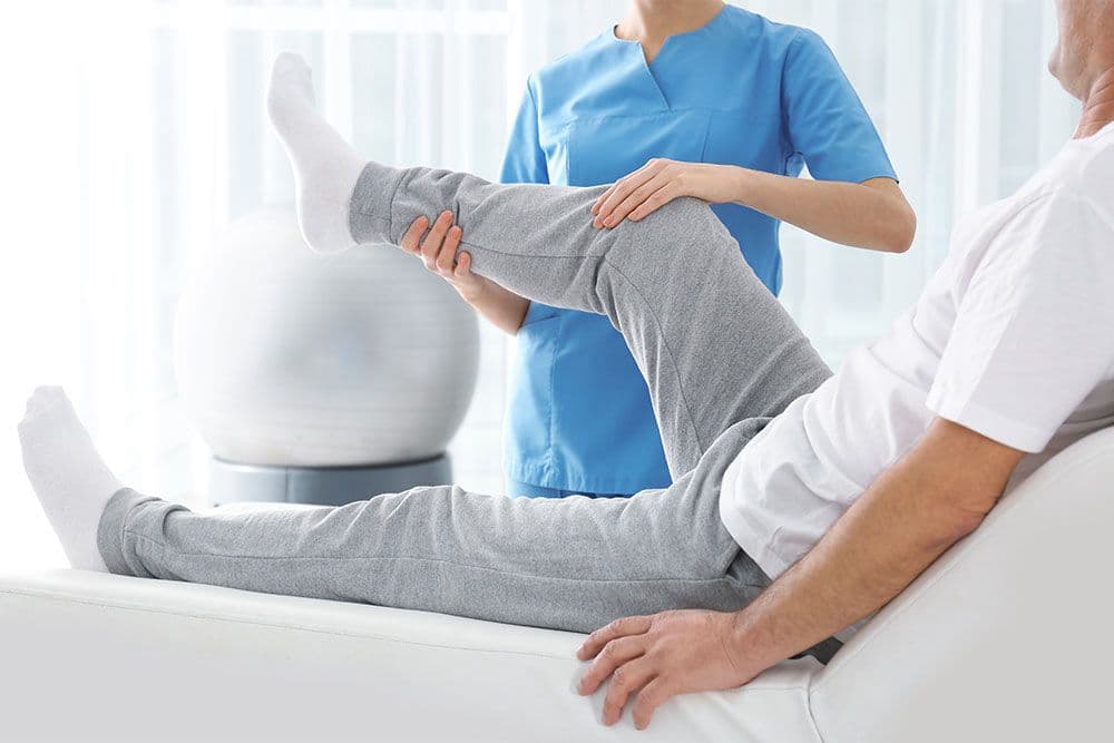 Physiotherapeut beugt das Knie eines liegenden Patienten während einer Mobilisationsübung, ein Gymnastikball steht daneben